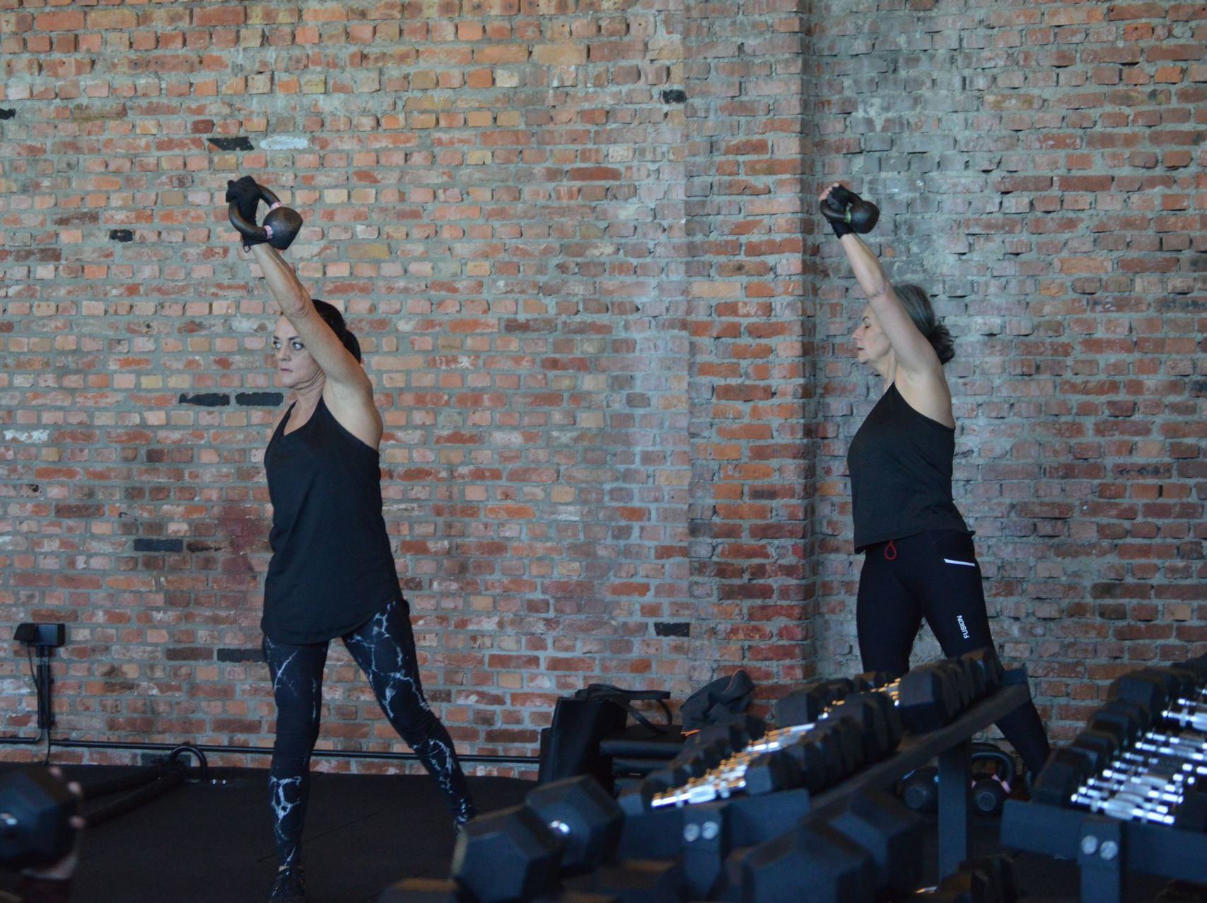 Een persoon tilt een kettlebell boven zijn hoofd in een fitness in hasselt met een bakstenen muur op de achtergrond.