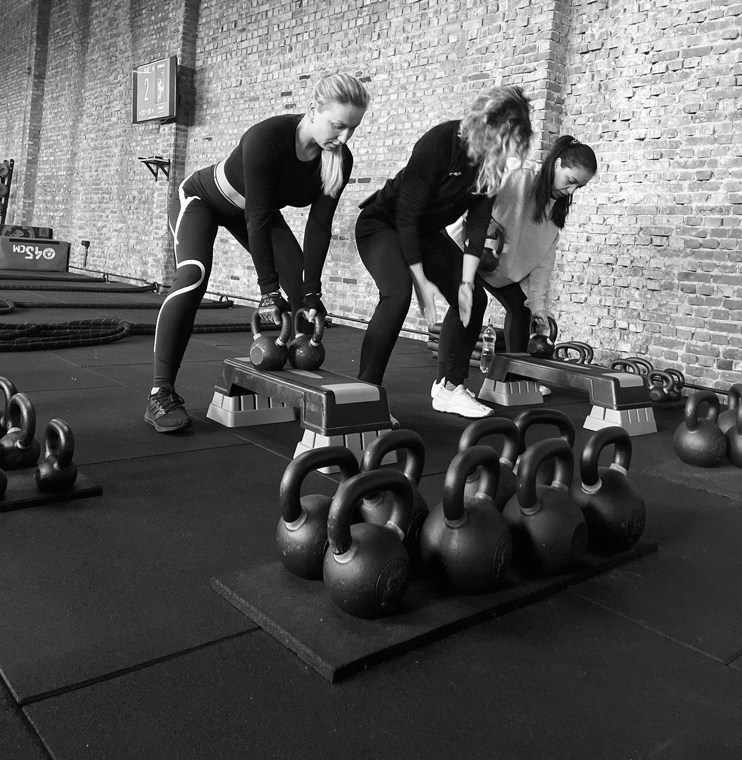 Drie mensen doen deadlifts met kettlebells in een sportschool met rijen kettlebells op de vloer.