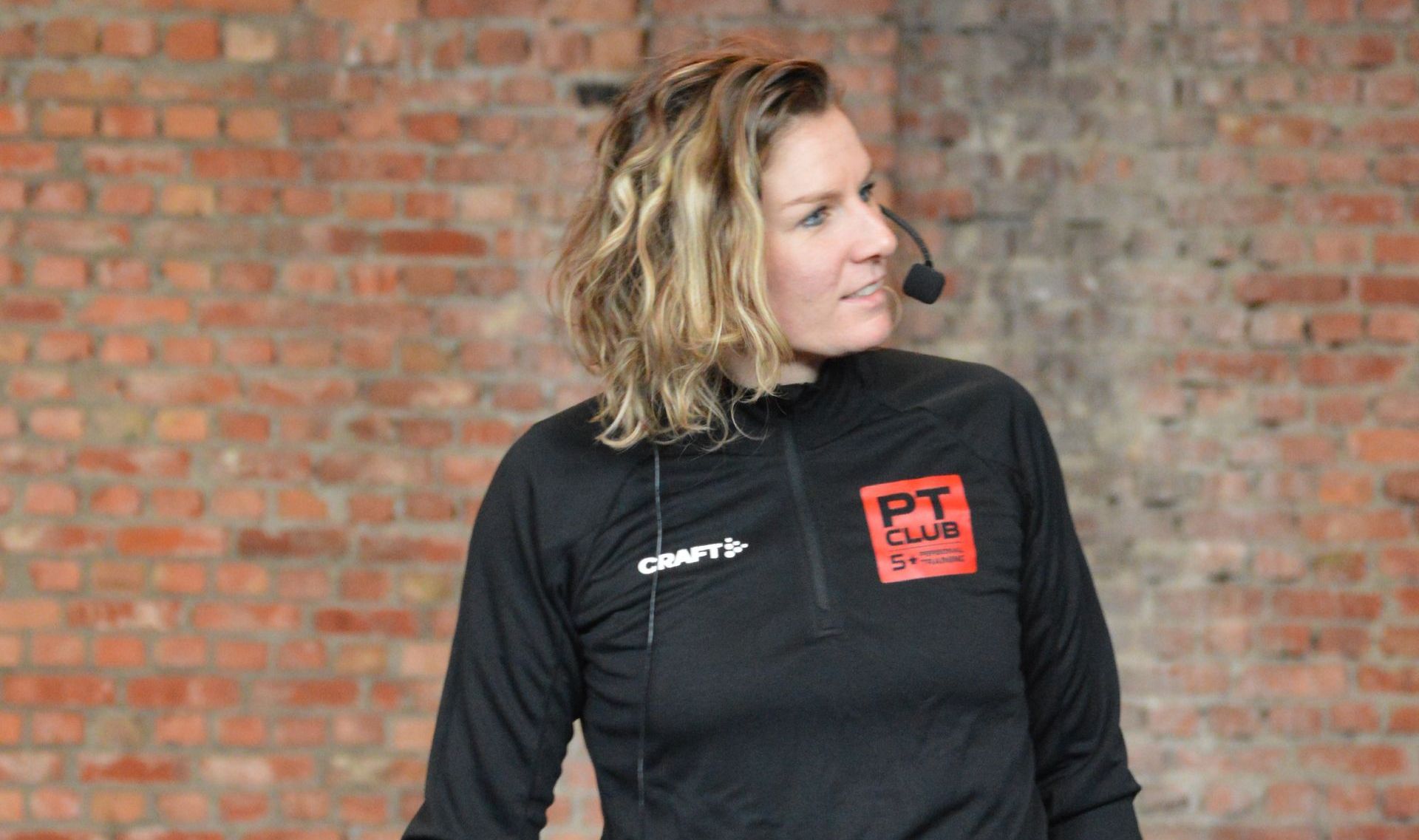 Vrouw in een zwarte jas met rood logo, kijkt opzij tegen een bakstenen muur.
