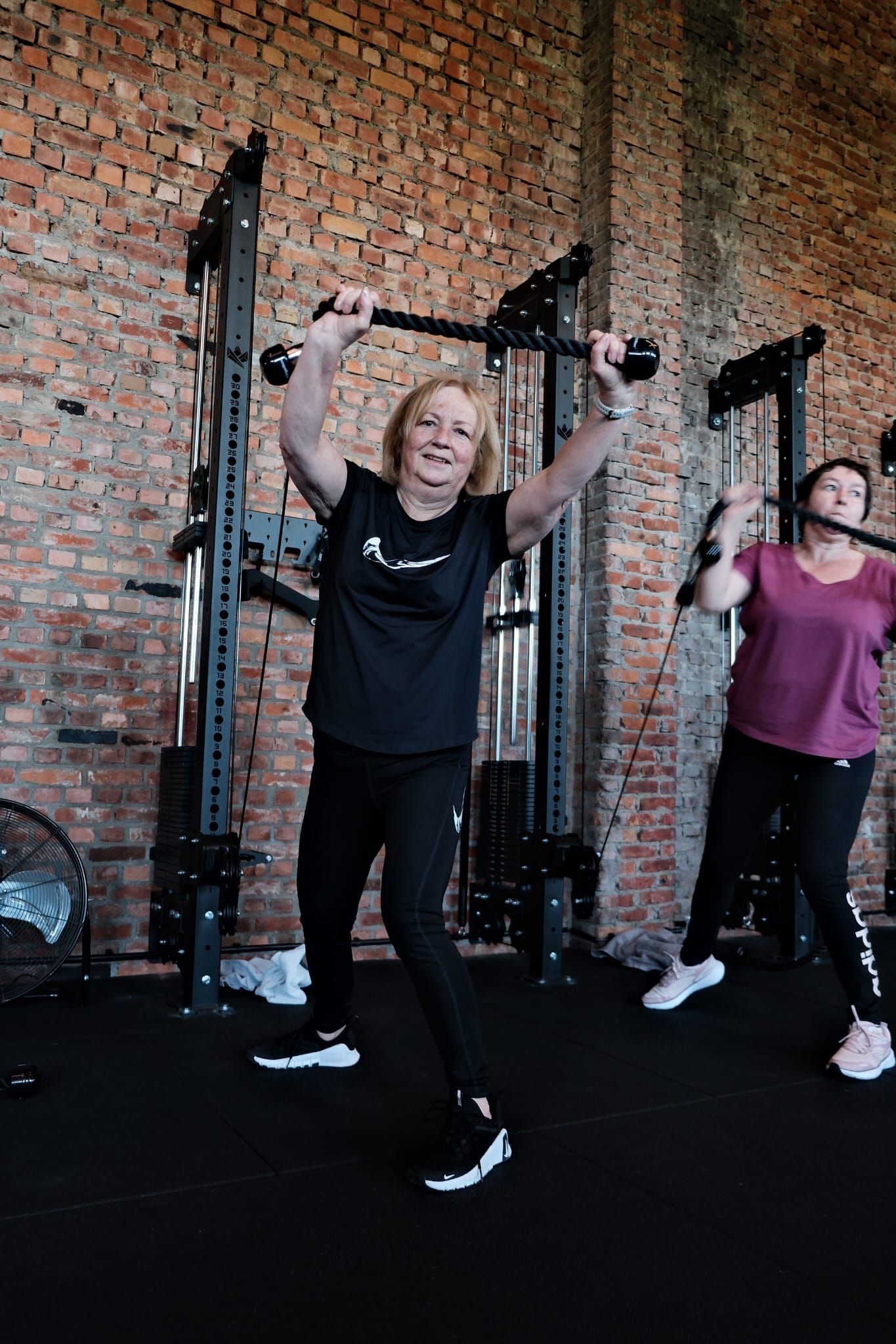 Twee mensen die in een sportschool met halters trainen, staand tegen een bakstenen muur.