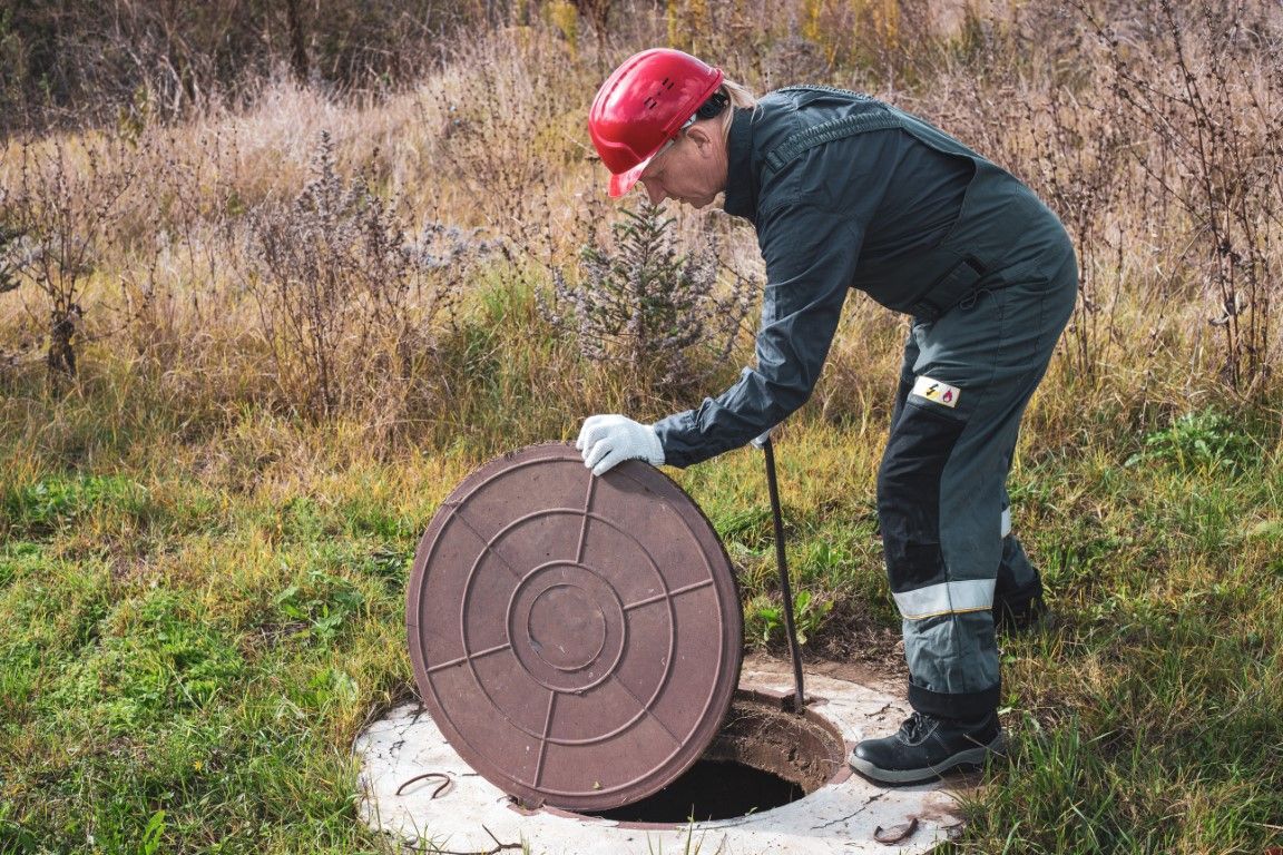 Person in red cap lifting a round manhole cover outdoors in a grassy field.