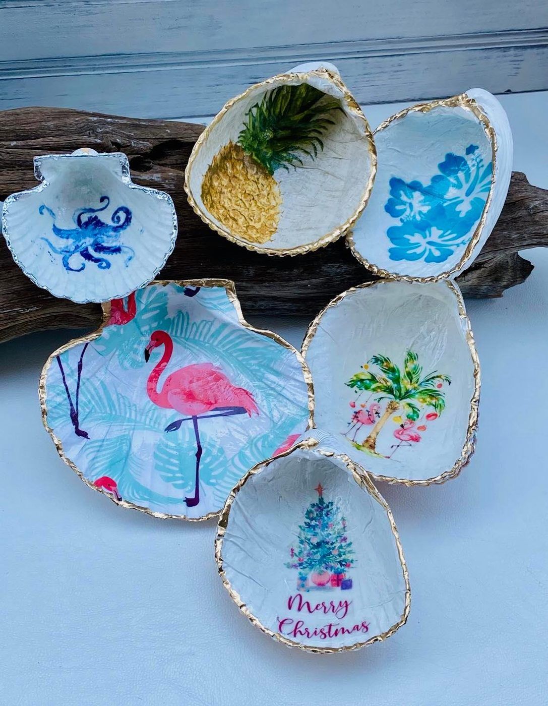 Hand-painted seashell ornaments with blue, pink, and floral designs on a rustic wood plank