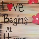 Handwritten “Love Begins At” with a red heart and pink flowers on a wood-look background