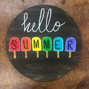 Round black sign with “hello summer” and colorful popsicle letters on a brown background