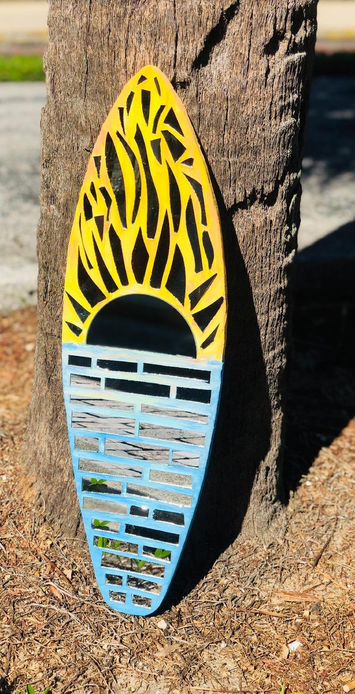 Colorful surfboard-shaped art leaning against a tree on sandy ground