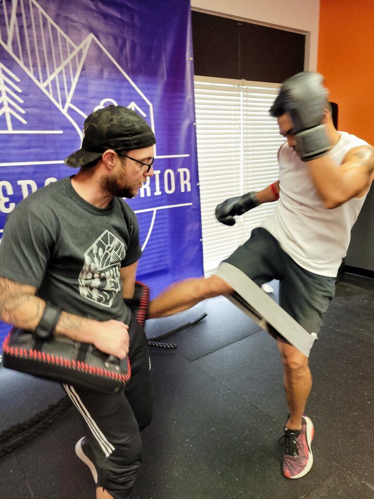 Two people spar in a gym, one kicking while the other blocks with a pad.