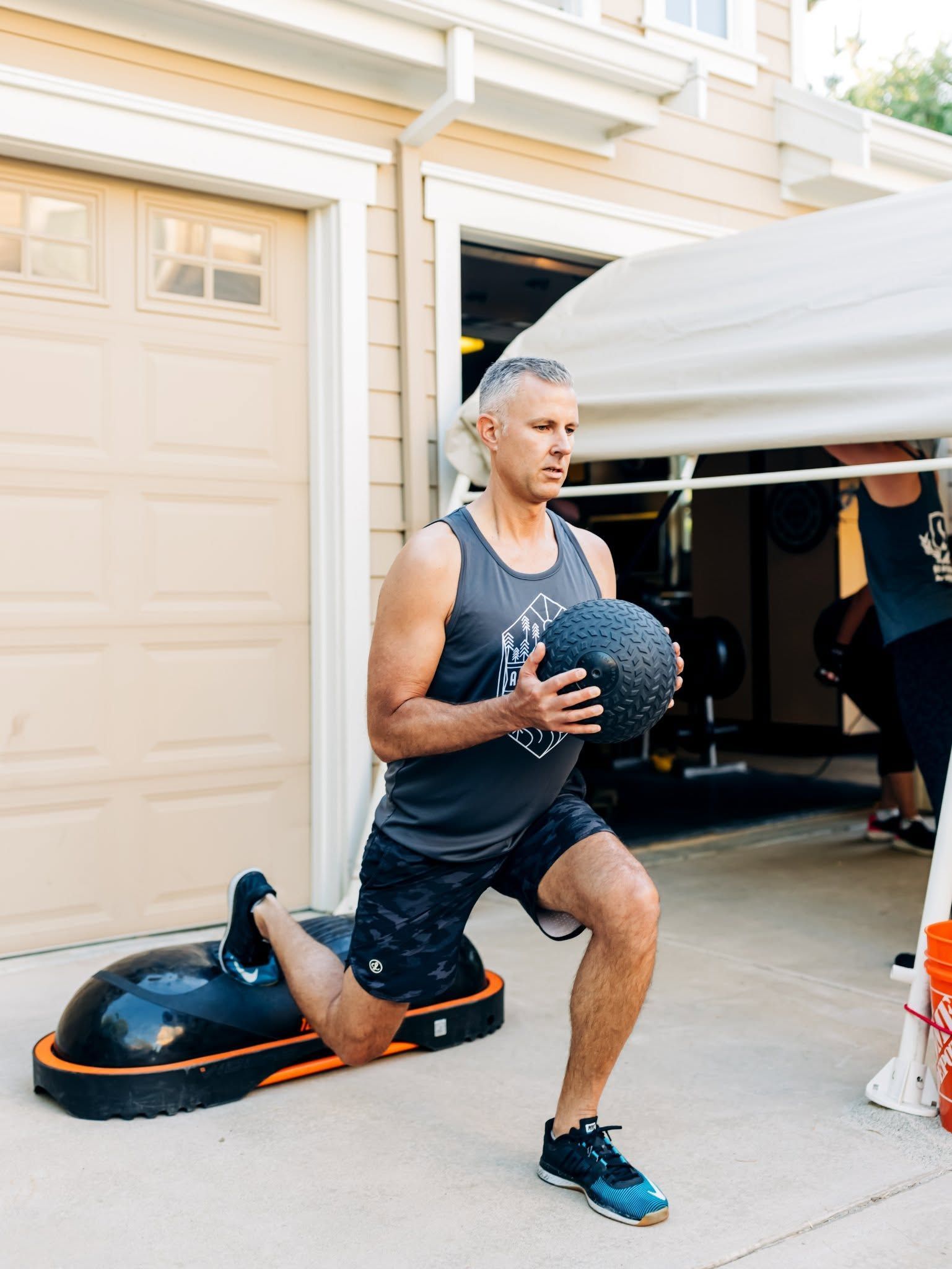 Person lunging with a medicine ball beside a garage, using a black balance trainer outdoors.