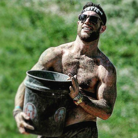 Smiling shirtless man carrying a large black bucket outdoors on grass