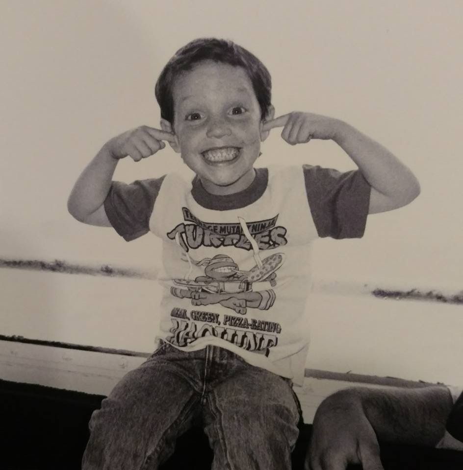 Smiling child sitting and pointing at their cheeks, wearing a graphic T-shirt.