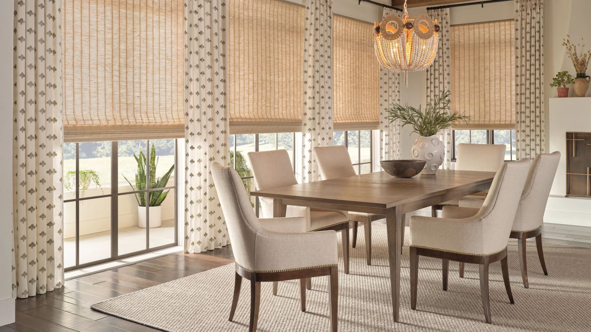 Dining room with a wooden table, chairs, and Hunter Douglas Provenance® Woven Wood Shades. Brown beams, chandelier, and patterned curtains.