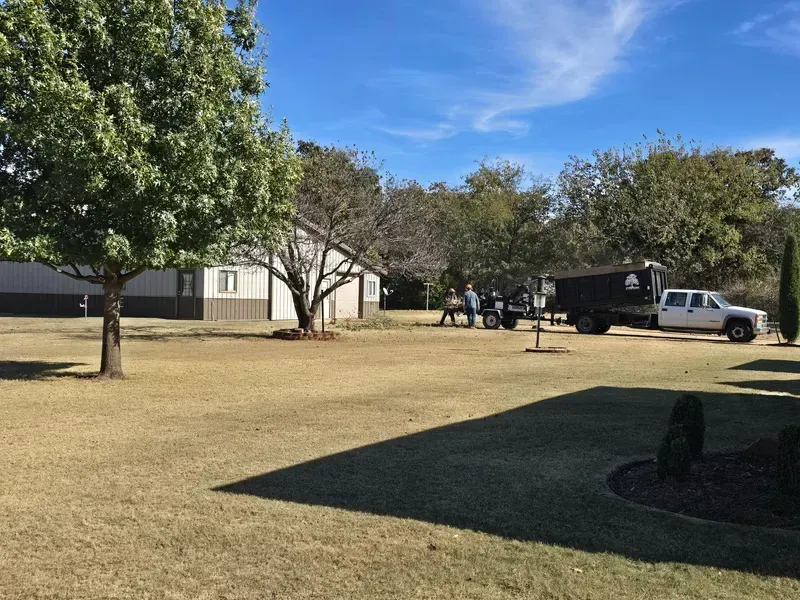 Suburban yard with trees, a house, and a white truck parked near a dark trailer on a sunny day