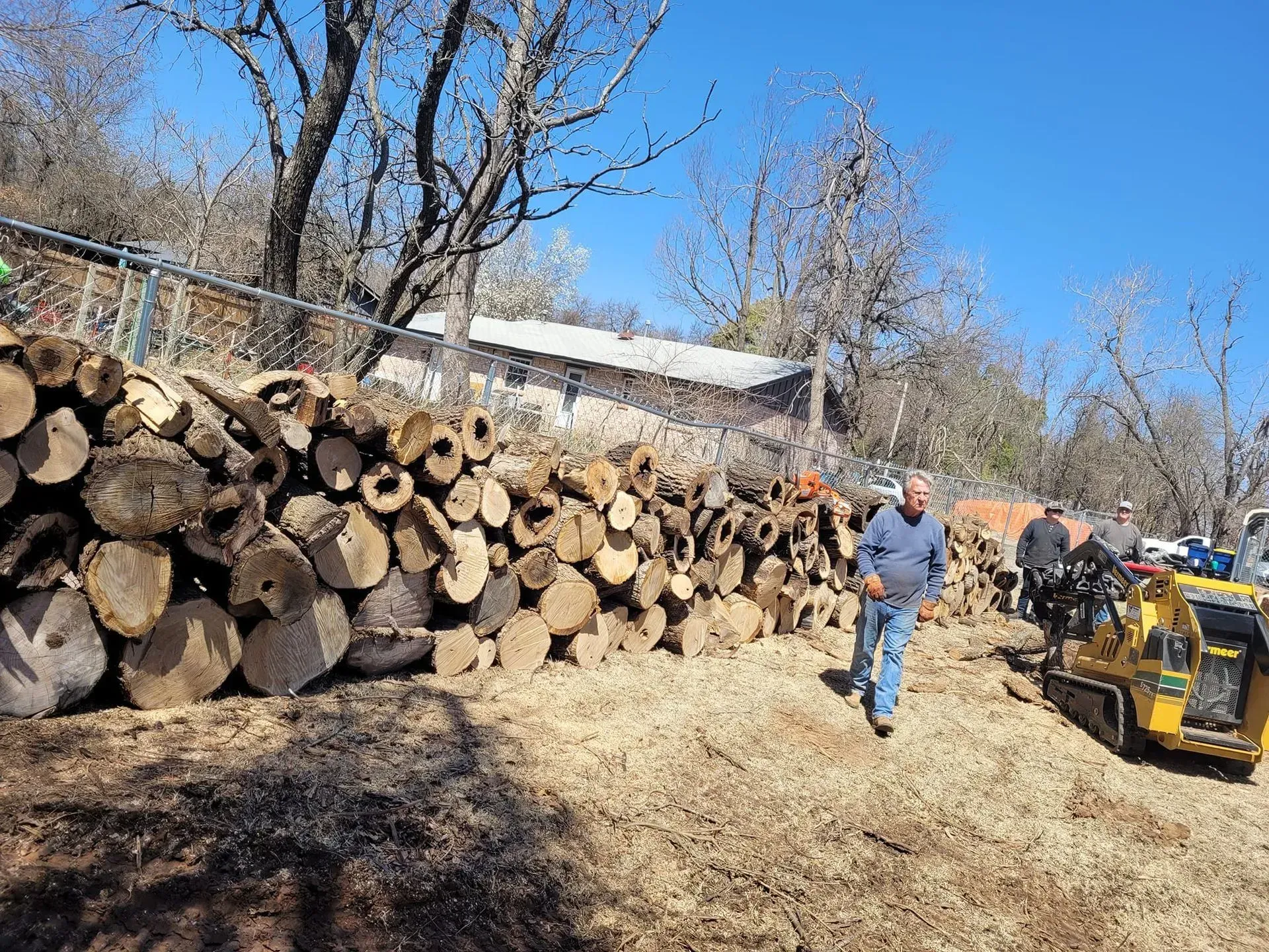 Man in blue stands beside a large stack of cut logs and a yellow loader on a sunny dirt road.