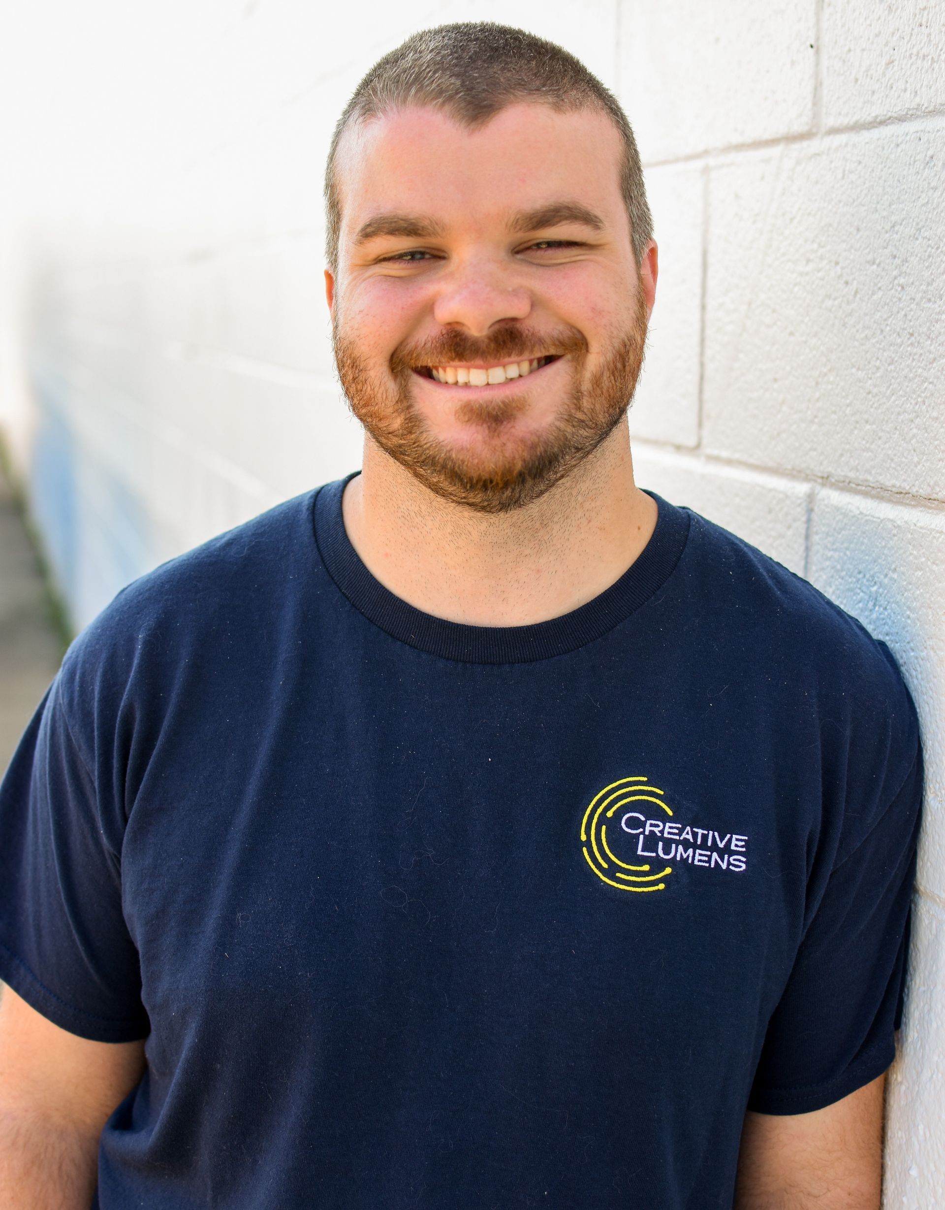 Man with short brown hair and beard smiles, leaning against a white brick wall. He wears a dark blue t-shirt with a logo.