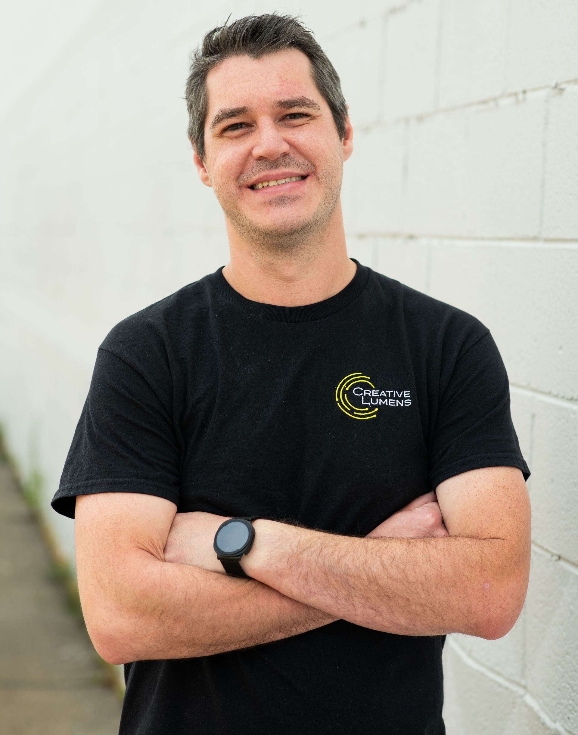 Man with glasses and black shirt smiling in front of a white brick wall.