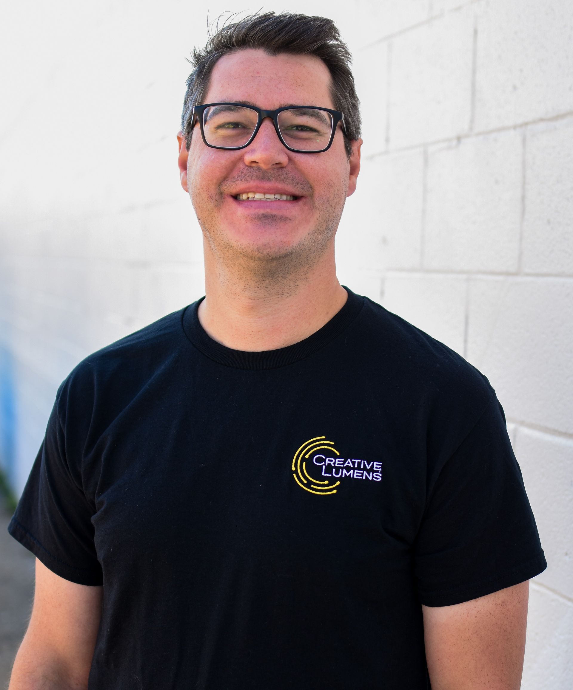 Man with glasses and black shirt smiling in front of a white brick wall.