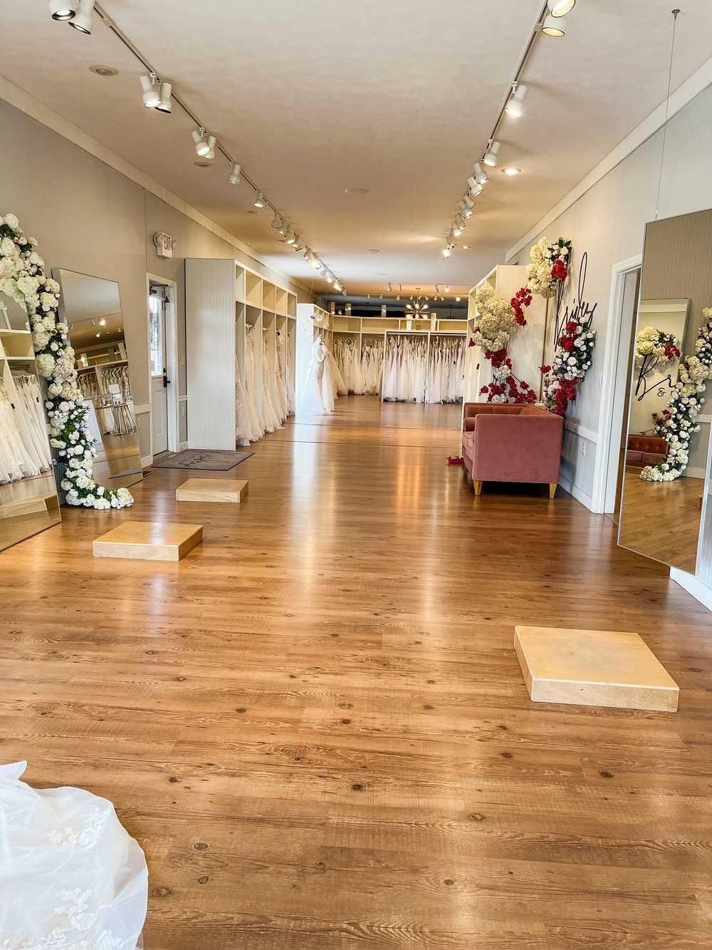 Interior of a bridal shop with dresses on display, mirrors, and a blush pink sofa. Wooden floors and white walls.
