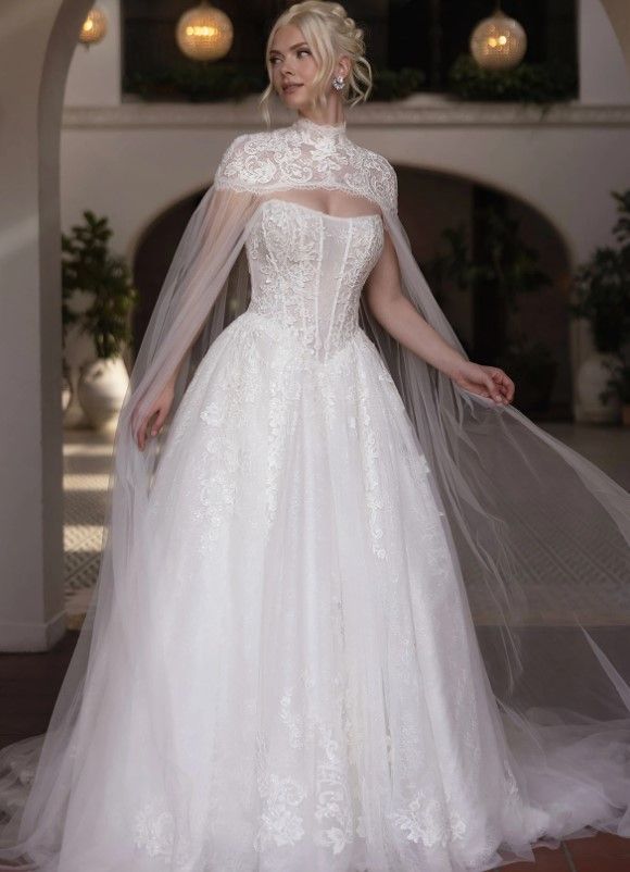 Woman in a white lace wedding gown with a sheer cape, standing in a softly lit interior courtyard.