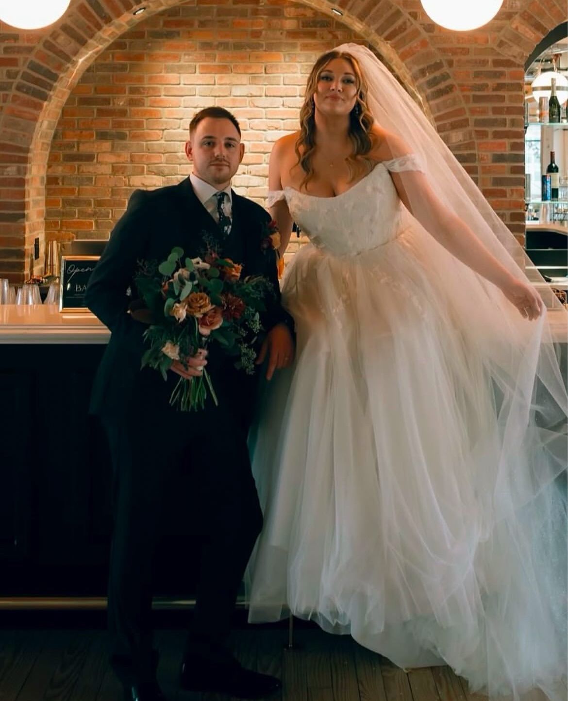 Bride and groom at a wedding. The bride in white dress, groom in black suit, posing indoors, smiling.