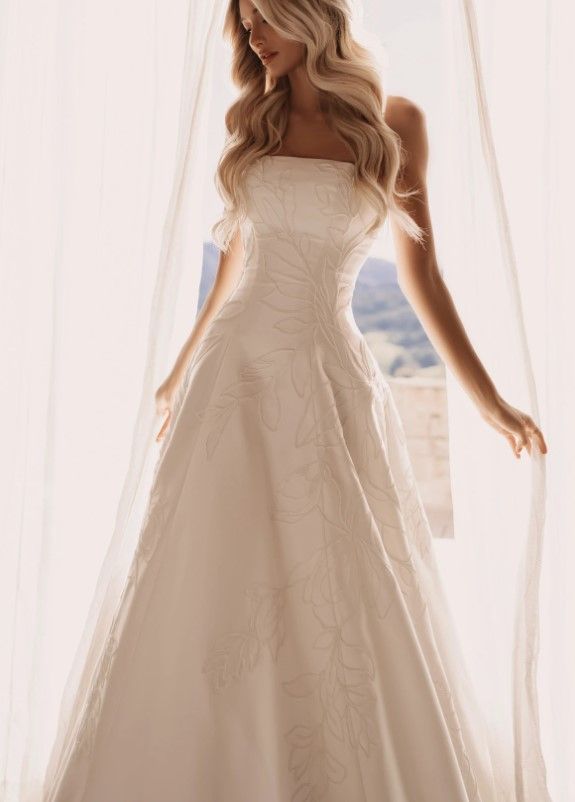 Strapless ivory wedding gown with a fitted bodice and flowing tulle skirt, worn indoors by a sheer curtain.