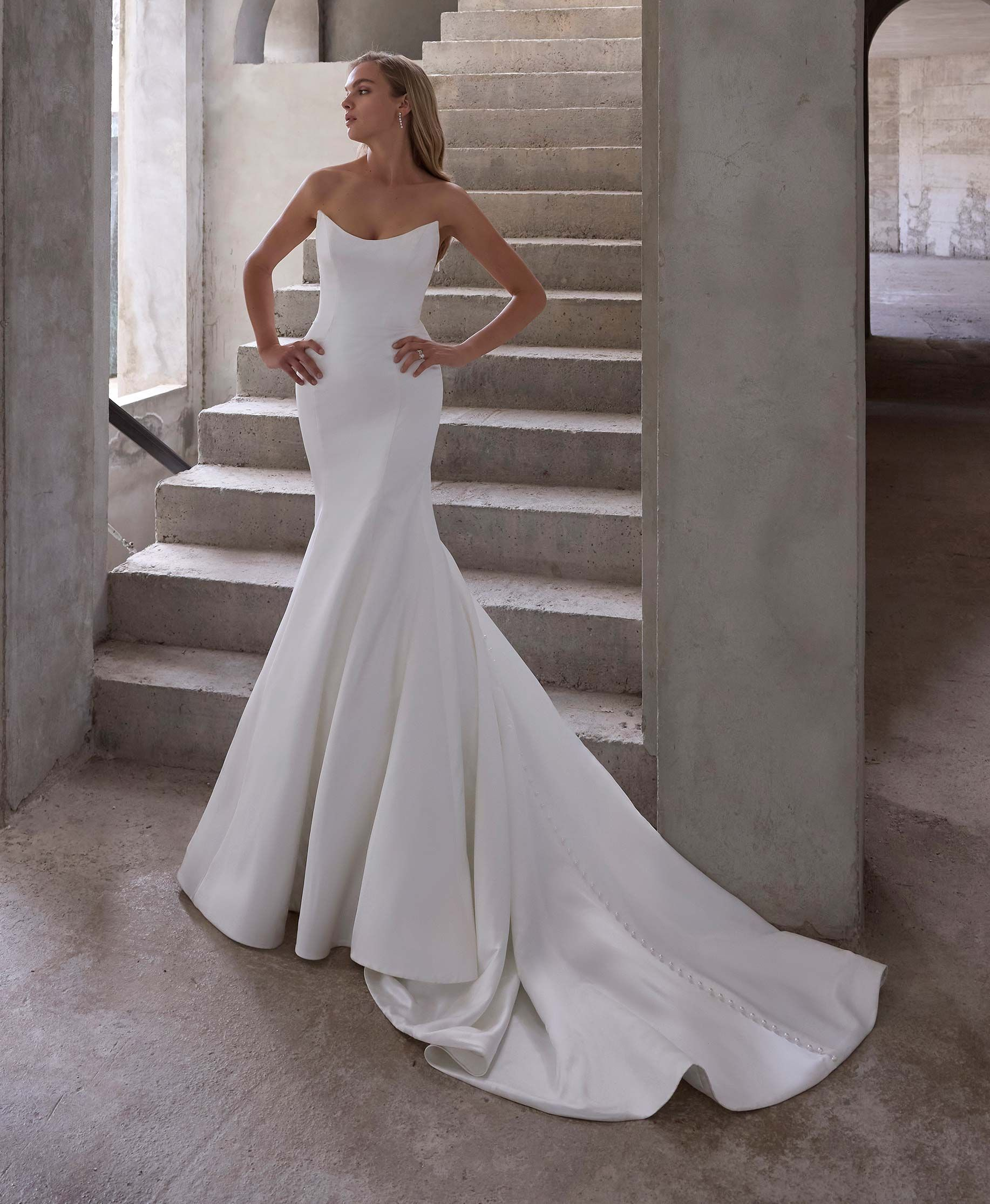 Bride in white strapless gown stands on stairs, hands on hips. Interior with arches, stone.