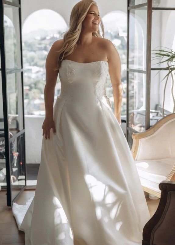 Woman in a white strapless gown standing by a sunlit window in a bright room