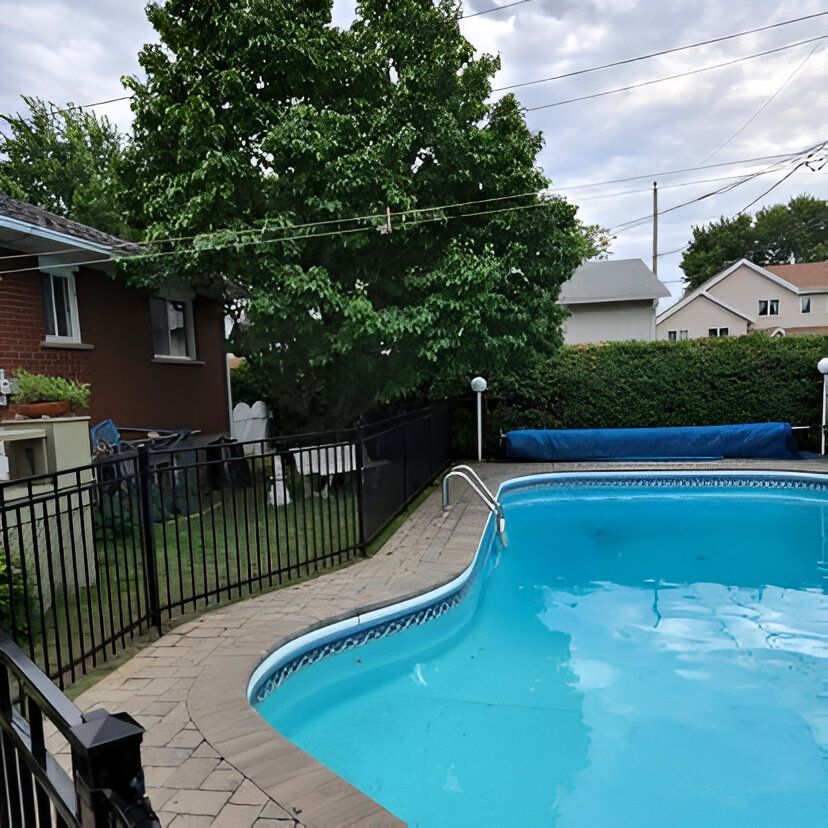 Backyard with a swimming pool surrounded by a fence and brick patio. Trees, houses, and an overcast sky are visible.