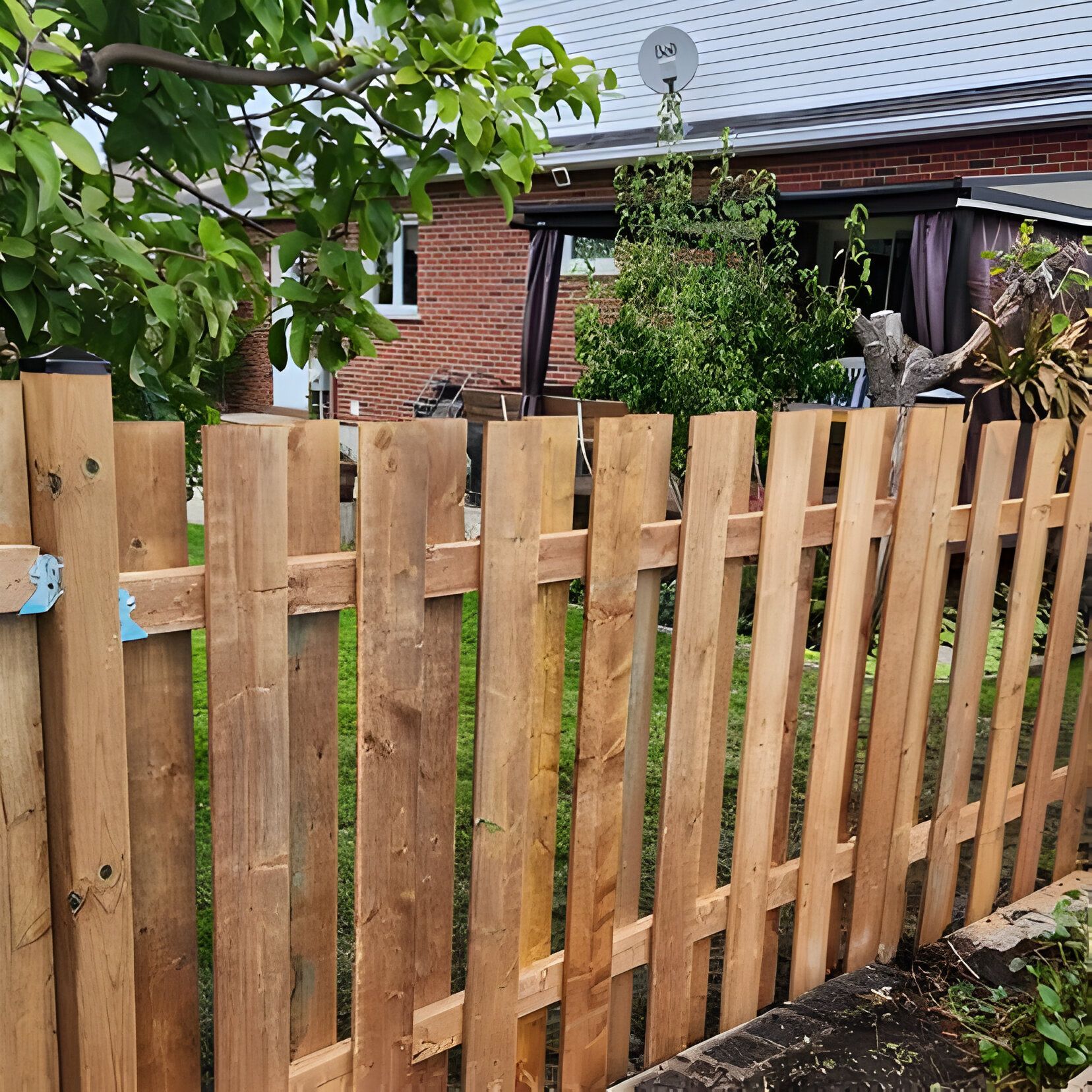 Wooden fence in front of a house, made of vertical planks, with trees and a building in the background.