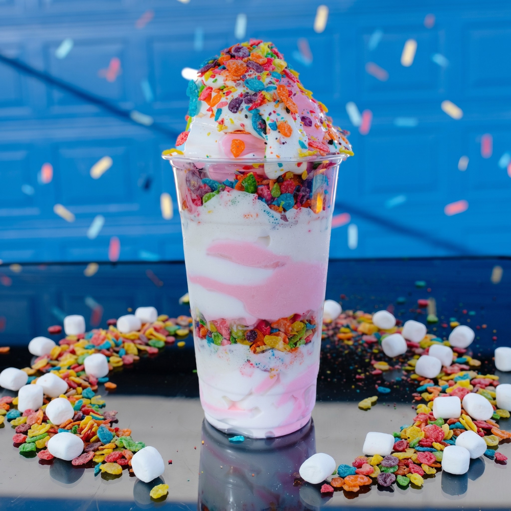 A clear plastic cup filled with pink and white swirled soft serve ice cream layered with colorful fruity cereal.