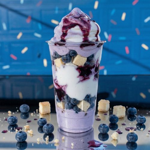 A blueberry cheesecake parfait with layers of cream, berry sauce, and cake cubes, topped with soft serve on a blue table.