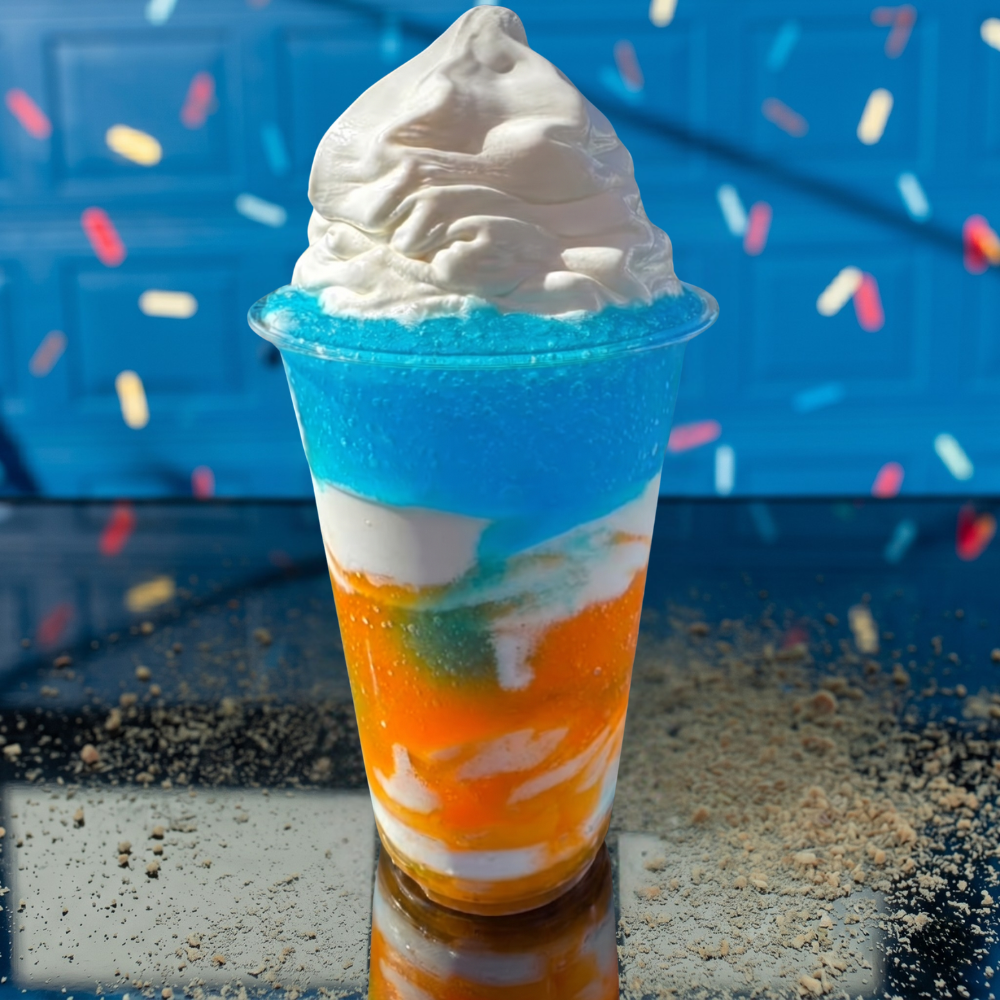 Colorful layered dessert in a clear cup with whipped cream on top, against a blue festive background.