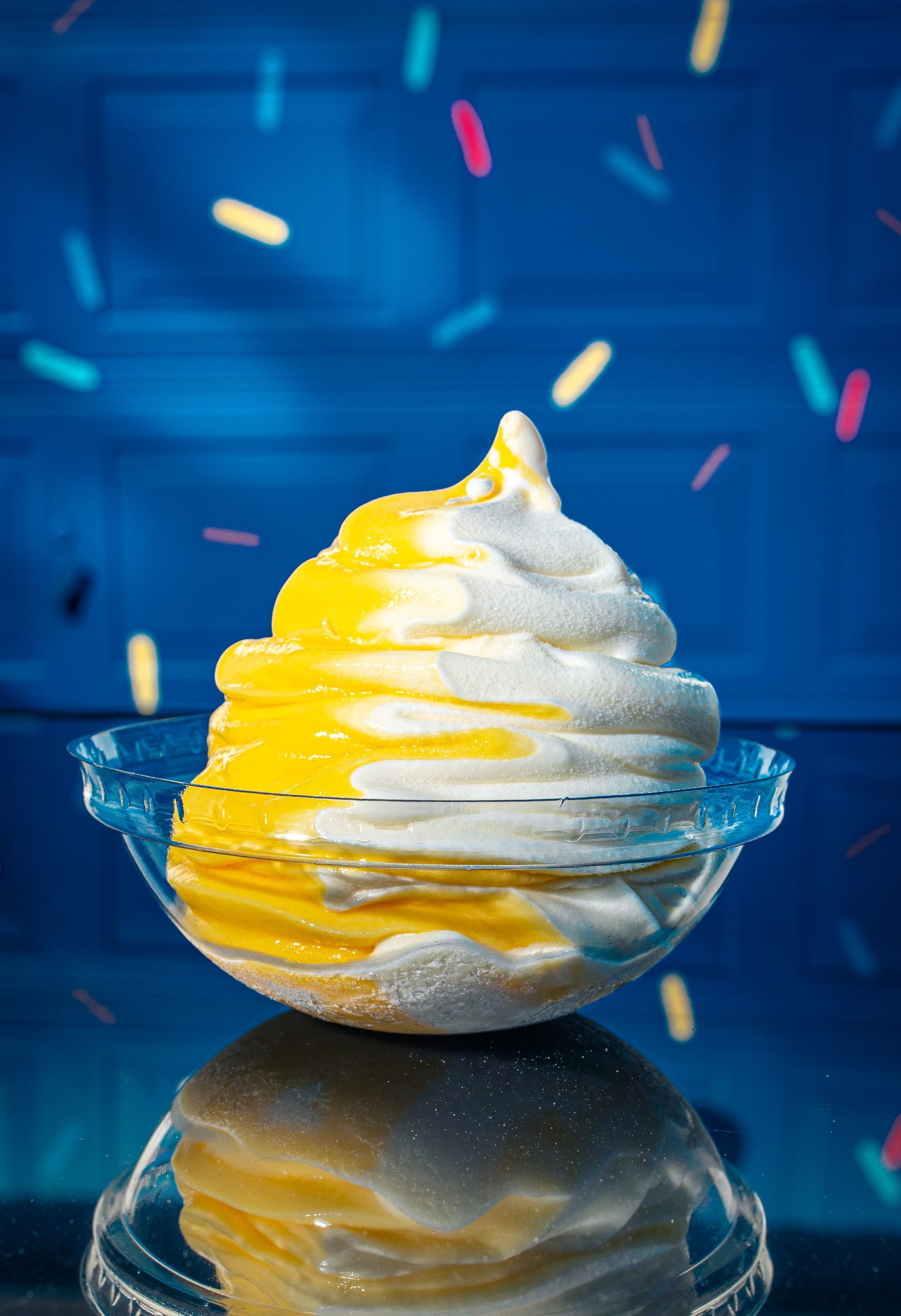 A swirl of yellow and white soft-serve ice cream in a small glass dish against a blue background with floating sprinkles.