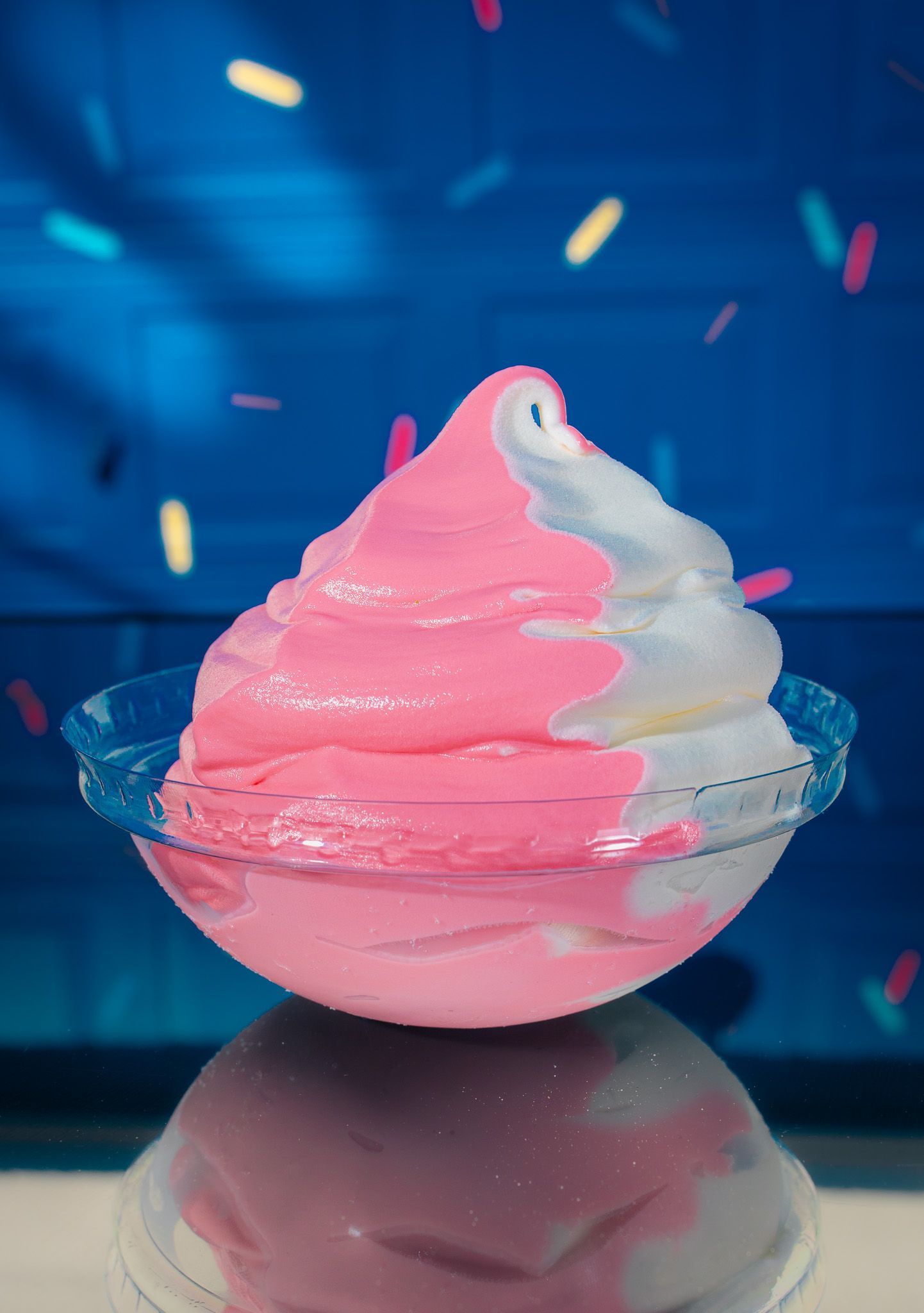 A swirling soft-serve treat with pink and white halves in a clear bowl against a blue background with floating sprinkles.