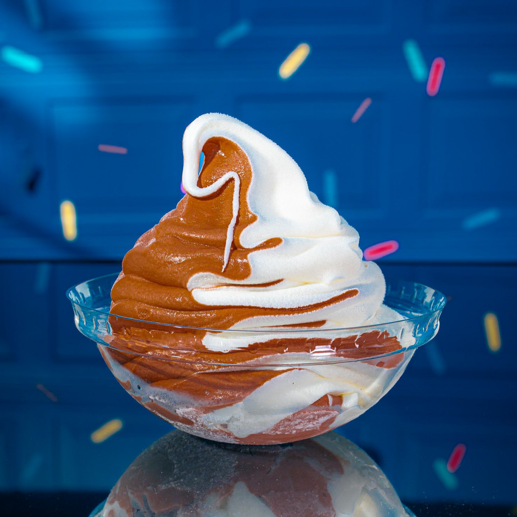 A swirl of chocolate and vanilla soft-serve ice cream in a clear bowl, set against a blue background with floating sprinkles.