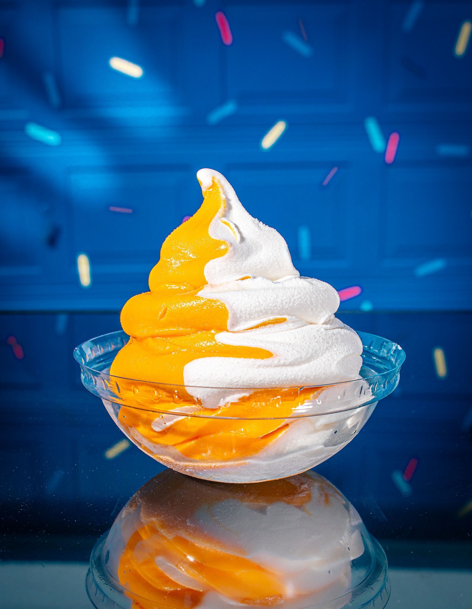 A swirl of orange and white soft-serve ice cream in a clear plastic bowl, set against a blue background with sprinkles.