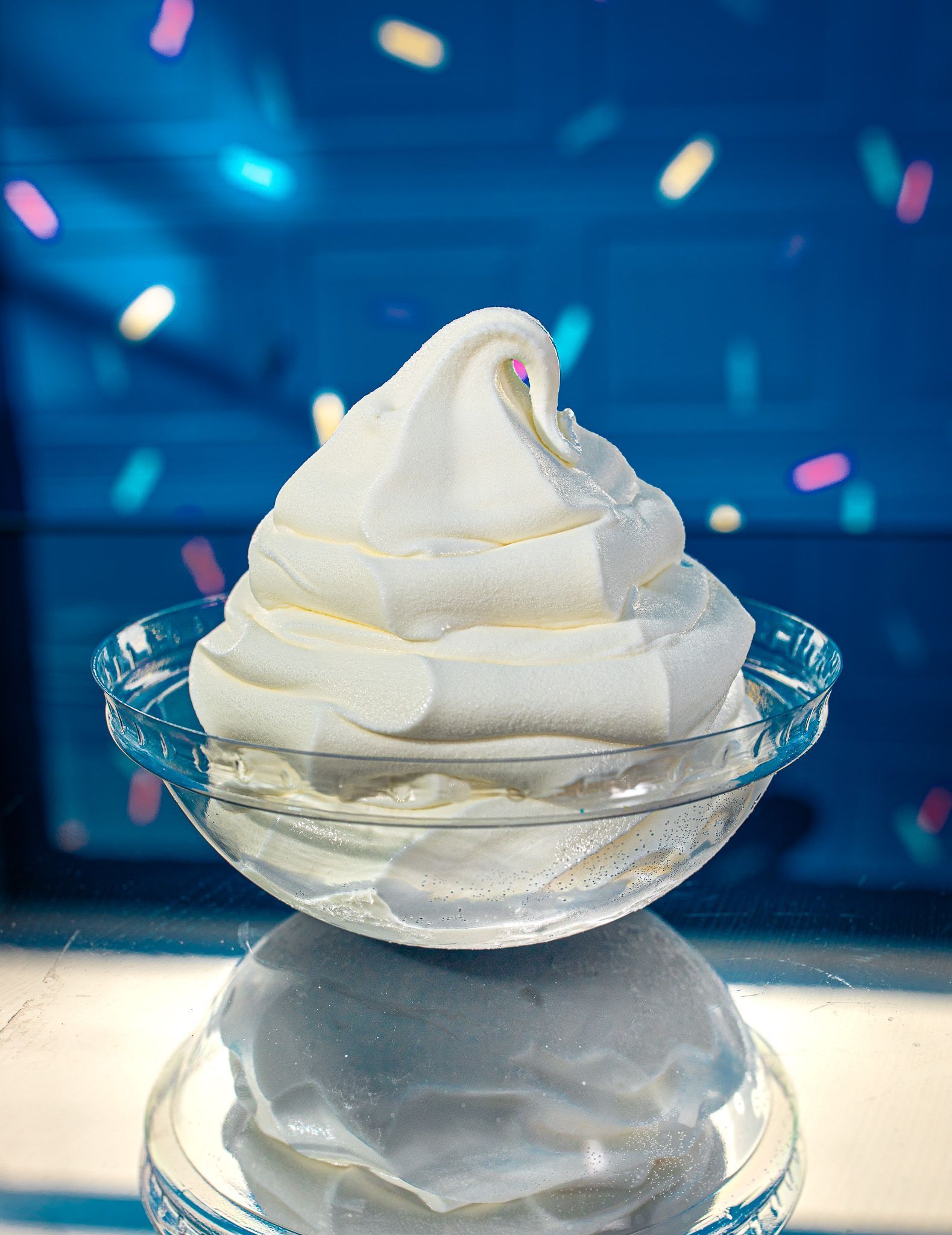 A swirl of white soft-serve ice cream in a clear glass bowl against a blue background with colorful floating sprinkles.