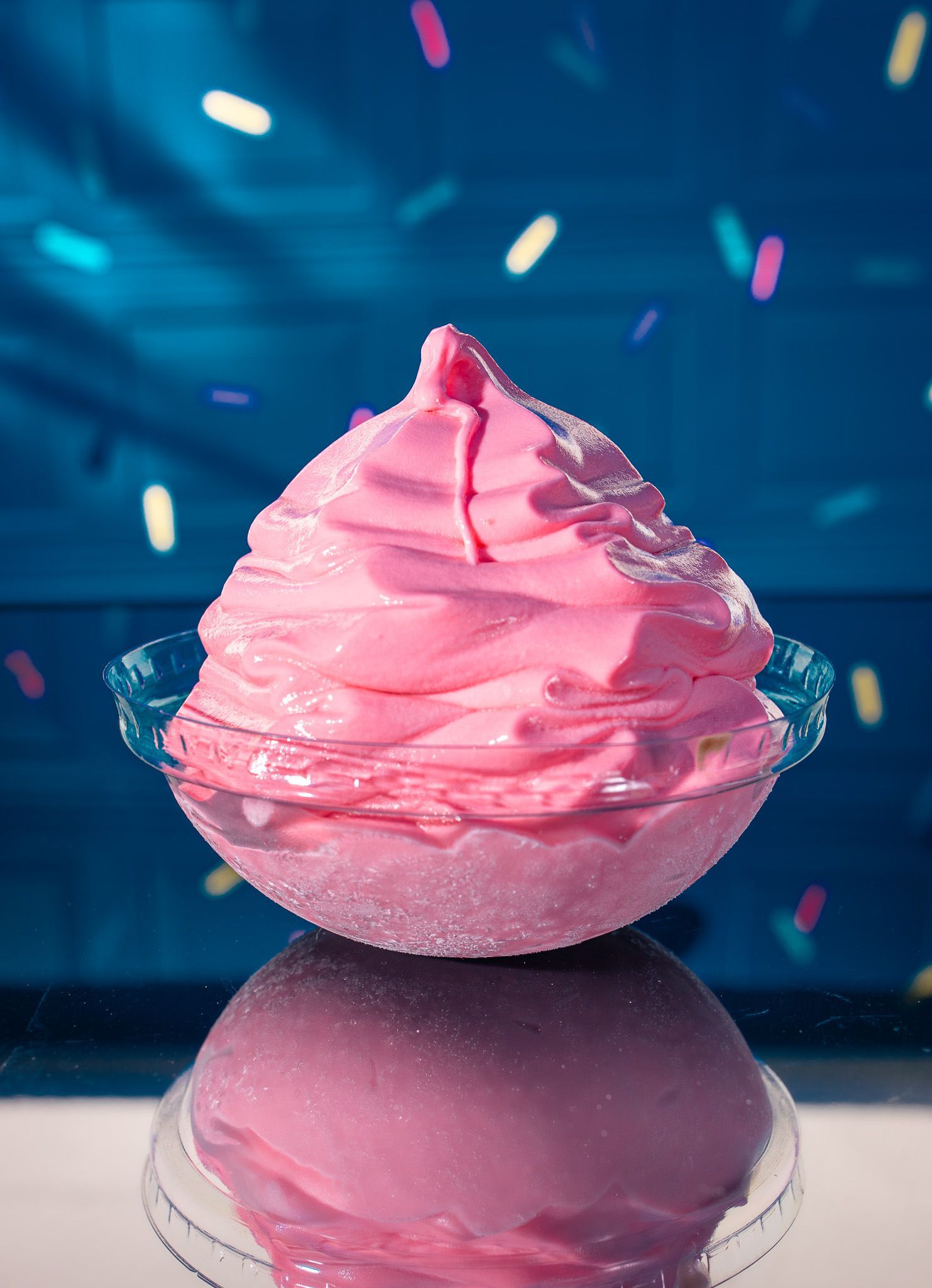 A swirl of bright pink soft-serve ice cream in a glass bowl set against a blue, confetti-dusted background.