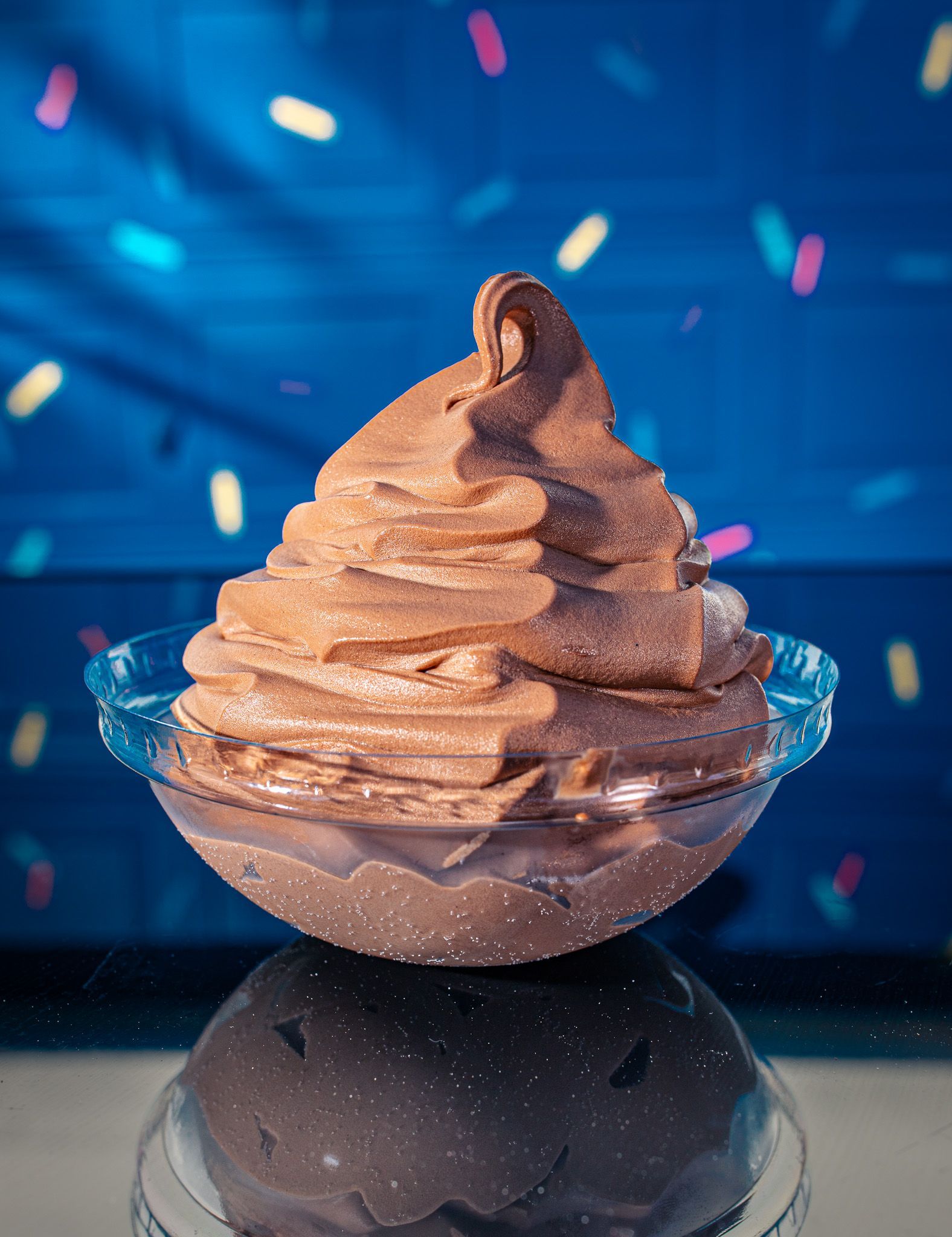 A swirl of chocolate soft-serve ice cream in a clear bowl, set against a blue background with colorful floating sprinkles.