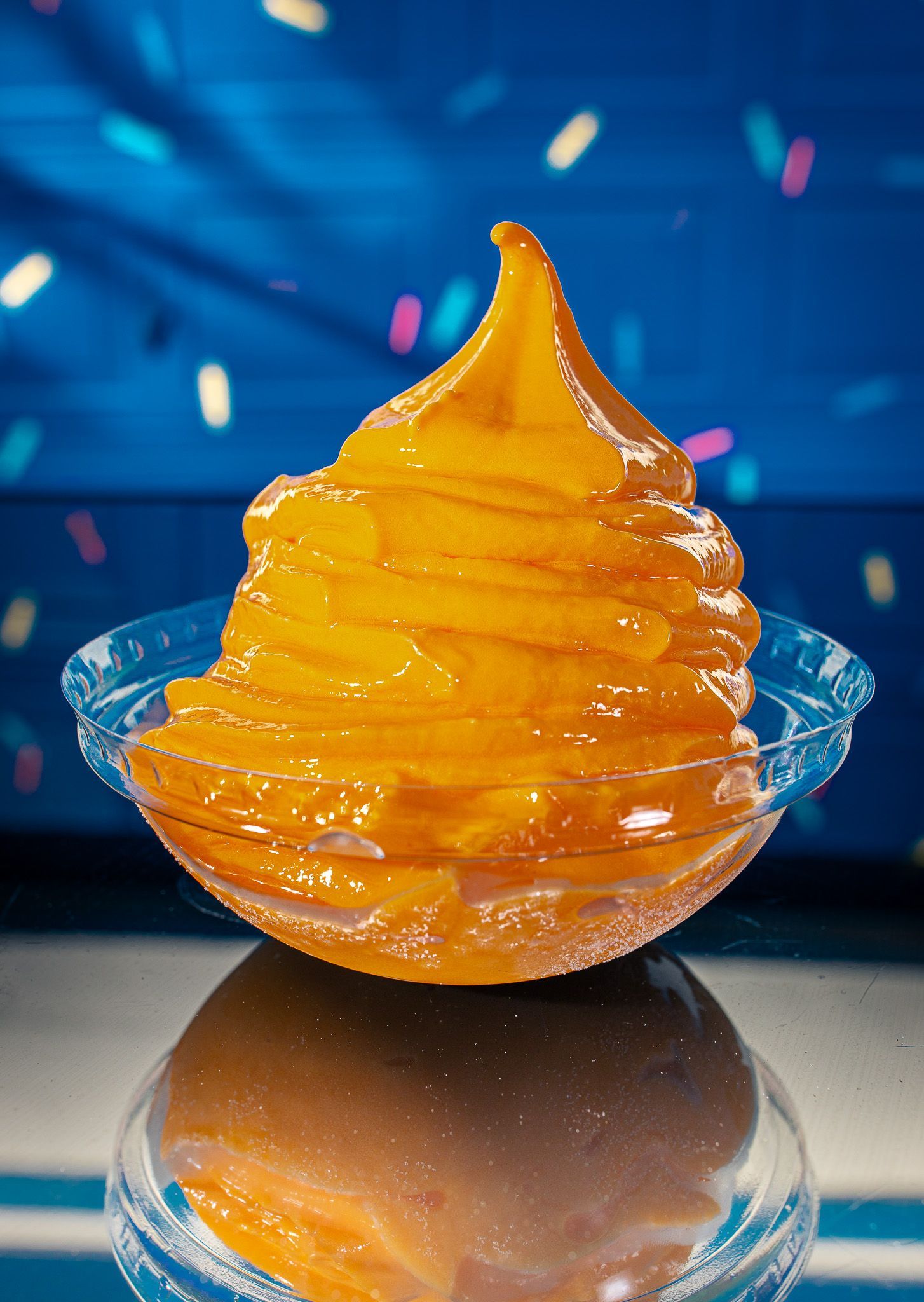 A swirl of orange soft-serve ice cream in a clear bowl, set against a blue background with colorful blurred confetti.