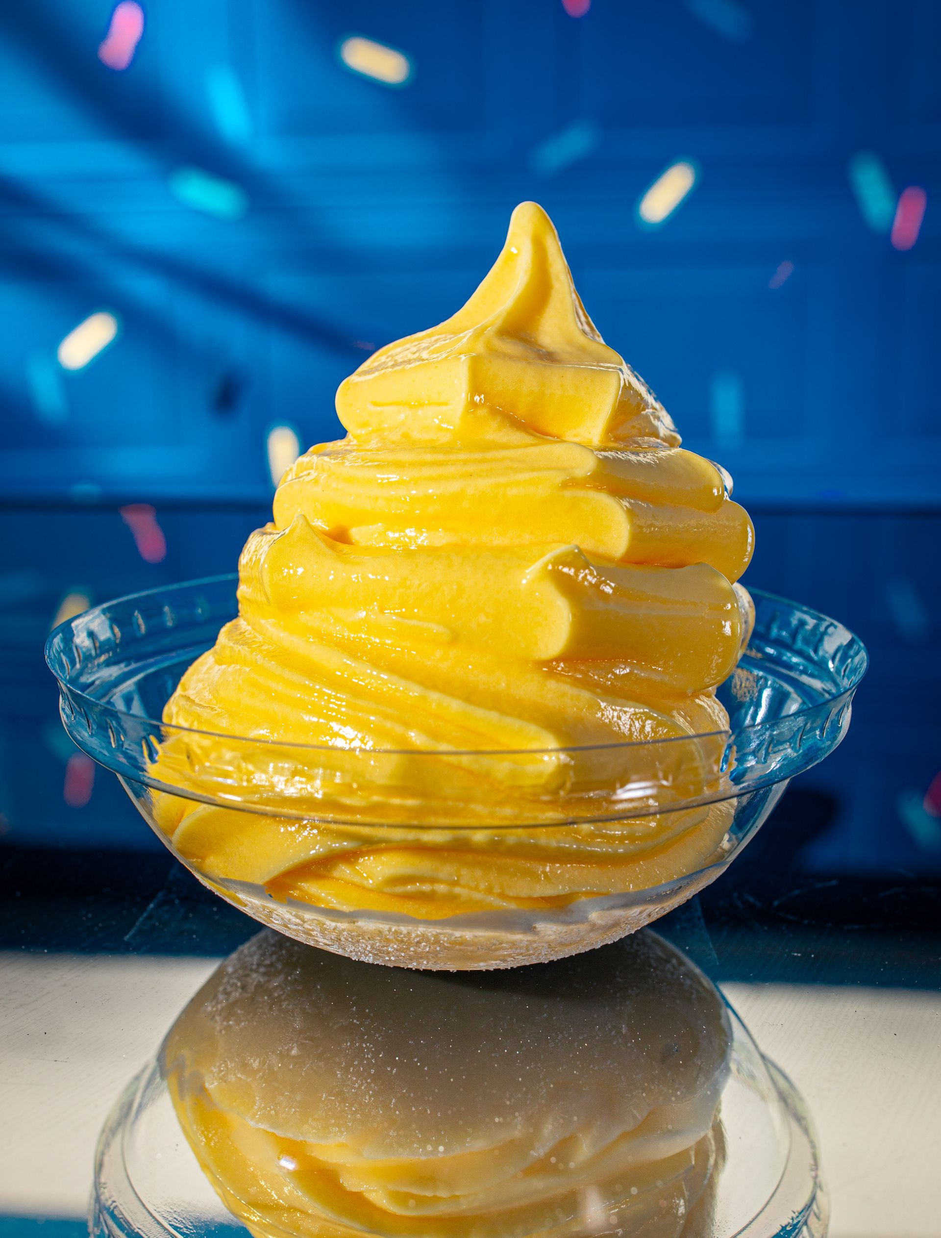 A swirl of yellow soft-serve ice cream in a clear glass bowl, reflected on a shiny surface against a blue backdrop.