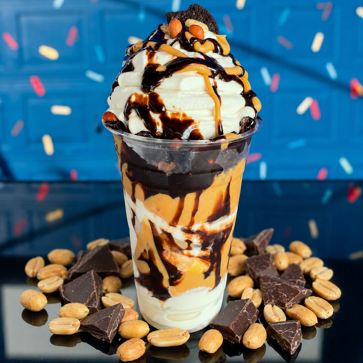 A clear plastic cup of layered vanilla ice cream, peanut butter, and chocolate sauce, topped with M&Ms, on a blue table.