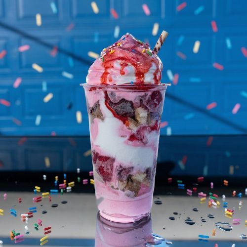 A strawberry sundae in a clear plastic cup with whipped cream, syrup, and a wafer stick against a blue background.