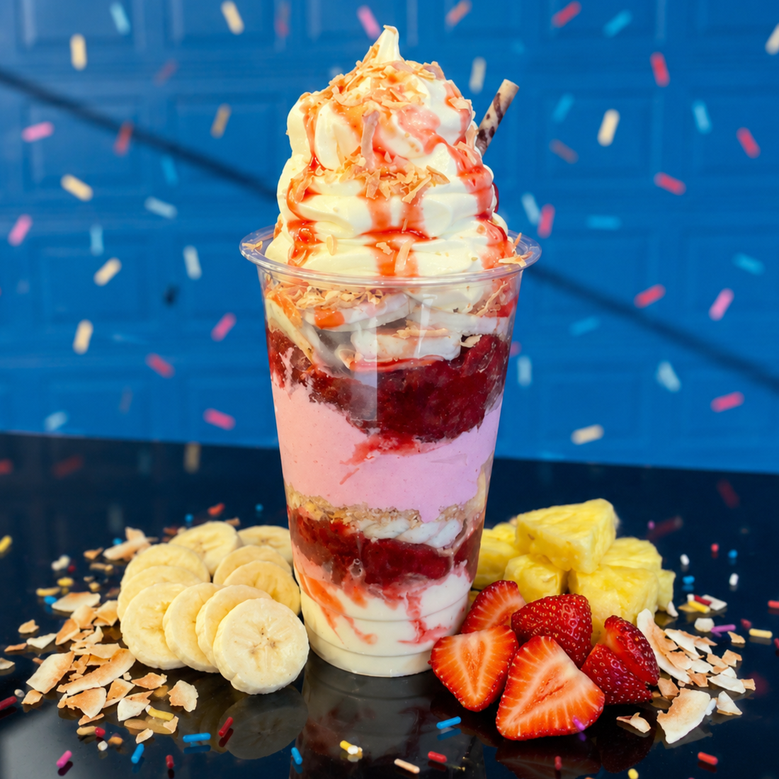 A layered dessert in a clear plastic cup topped with whipped cream, surrounded by banana slices, pineapple, and berries.