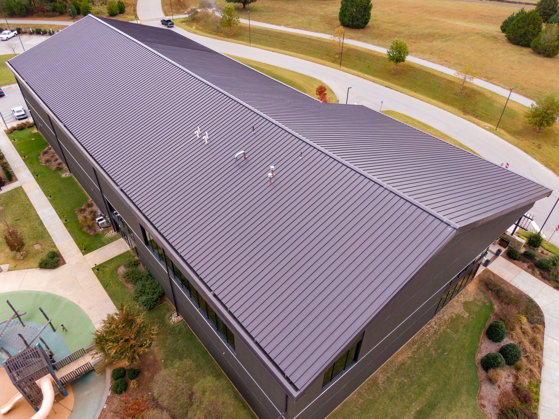 Aerial view of a rectangular, dark metal-roofed building next to a park and walkway in a suburban setting.