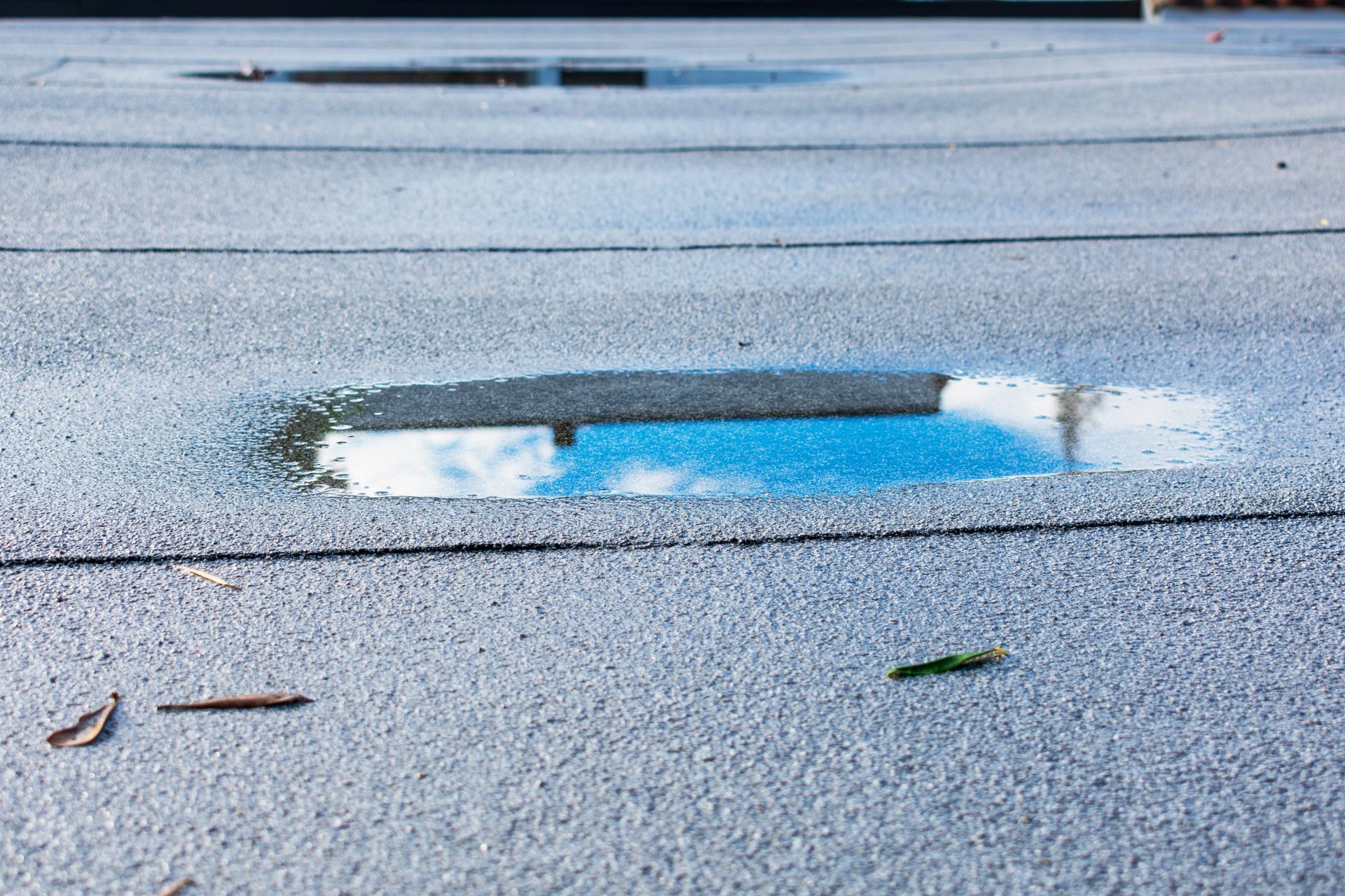 A close-up view of two puddles on a flat, gray, textured roof membrane reflecting the blue sky and clouds.