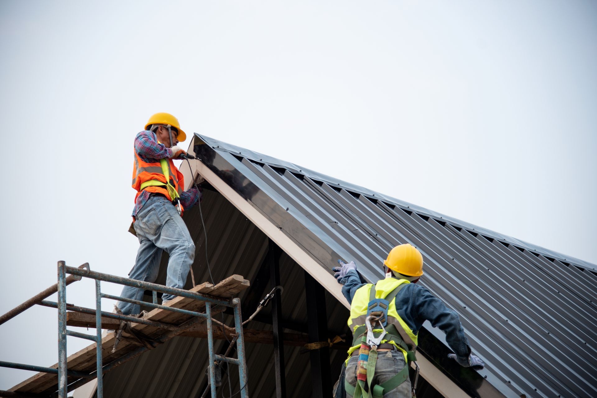 Two workers in hard hats and safety harnesses stand on scaffolding and a sloped roof, installing dark metal roofing panels.