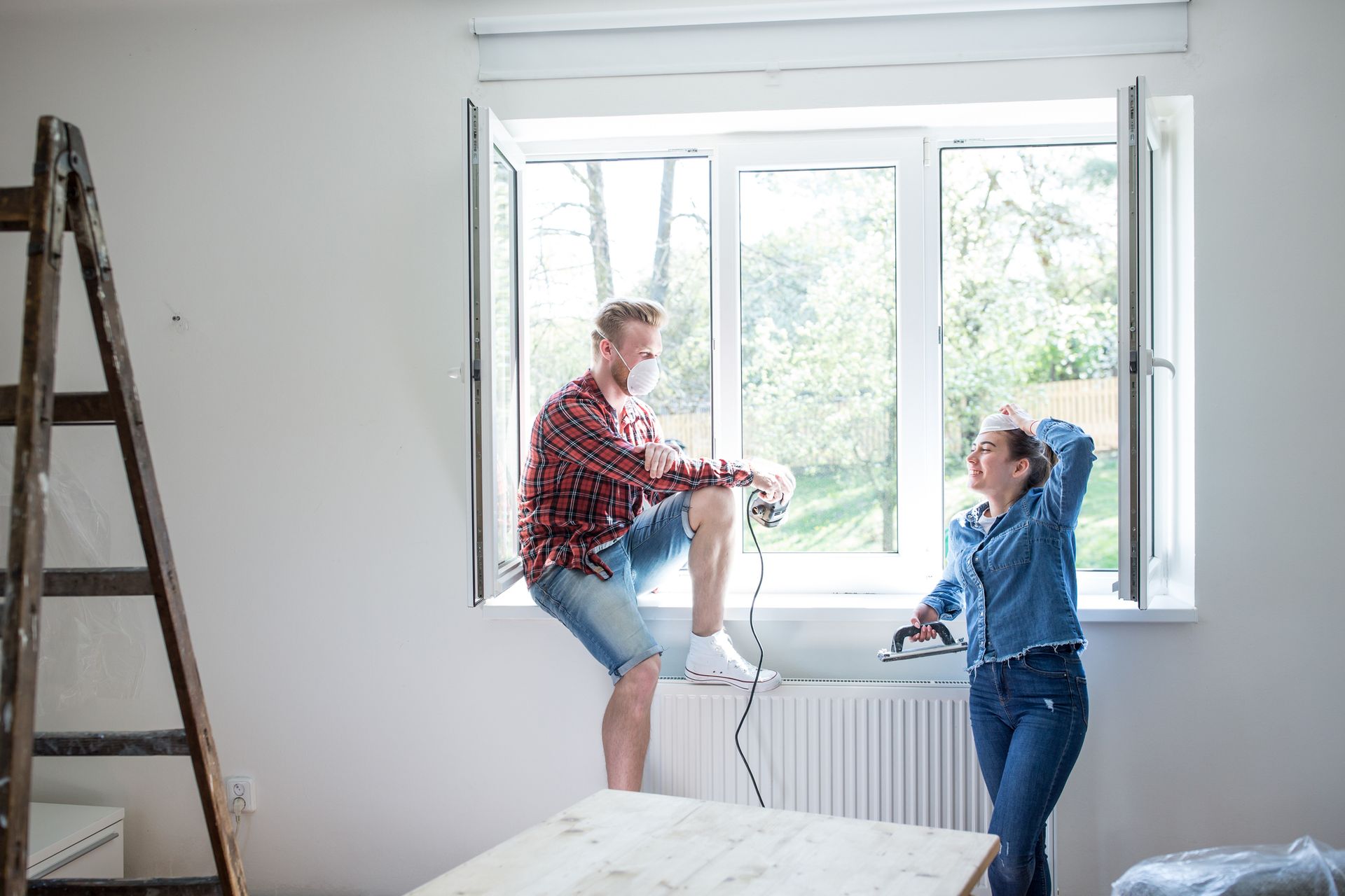 Two people replacing a window during a home renovation, ladder and tools visible. Two people replacing a window during a home renovation, ladder and tools visible.