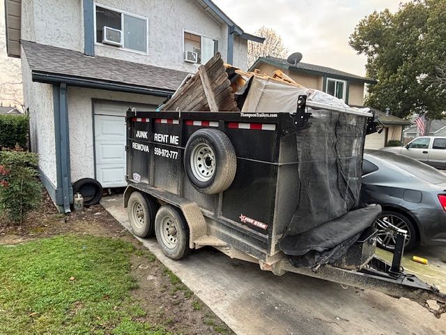 A black junk removal trailer filled with debris parked in a residential driveway in front of a house.