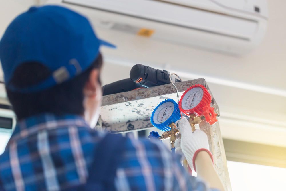 Technician installing a wall-mounted air conditioner with pressure gauges attached on a ladder