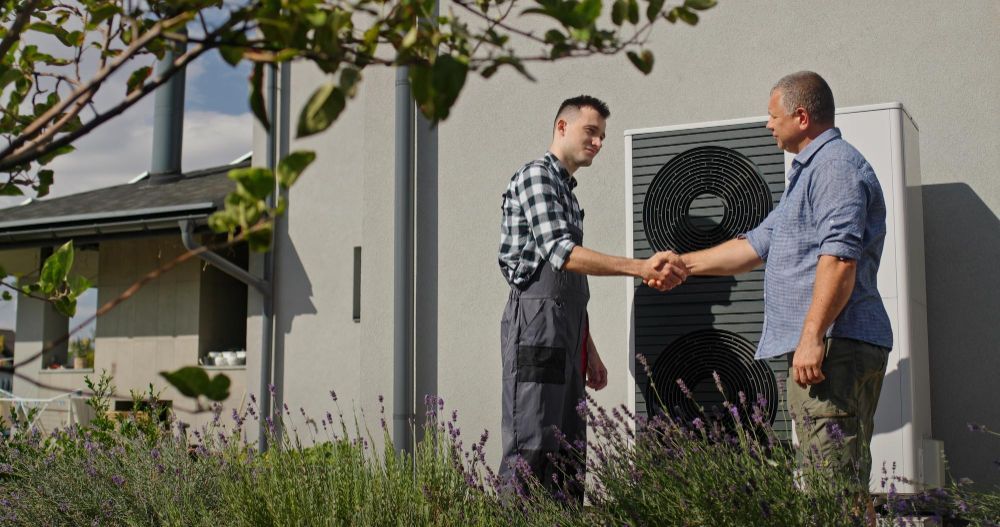 Two men shake hands beside an outdoor HVAC unit outside a house.