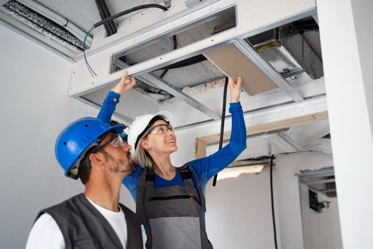Two workers inspecting ceiling ductwork in a white room, one in a blue hard hat pointing upward.